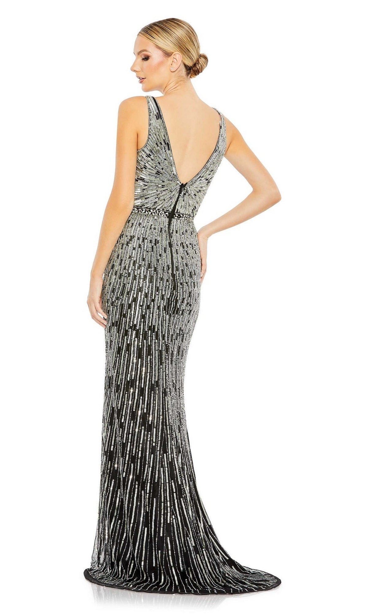 A woman stands facing away, wearing the Long Formal Dress 5483 by Mac Duggal—a black, floor-length, sleeveless fitted gown with silver sequin patterns, a deep V-back, and a flattering silhouette.